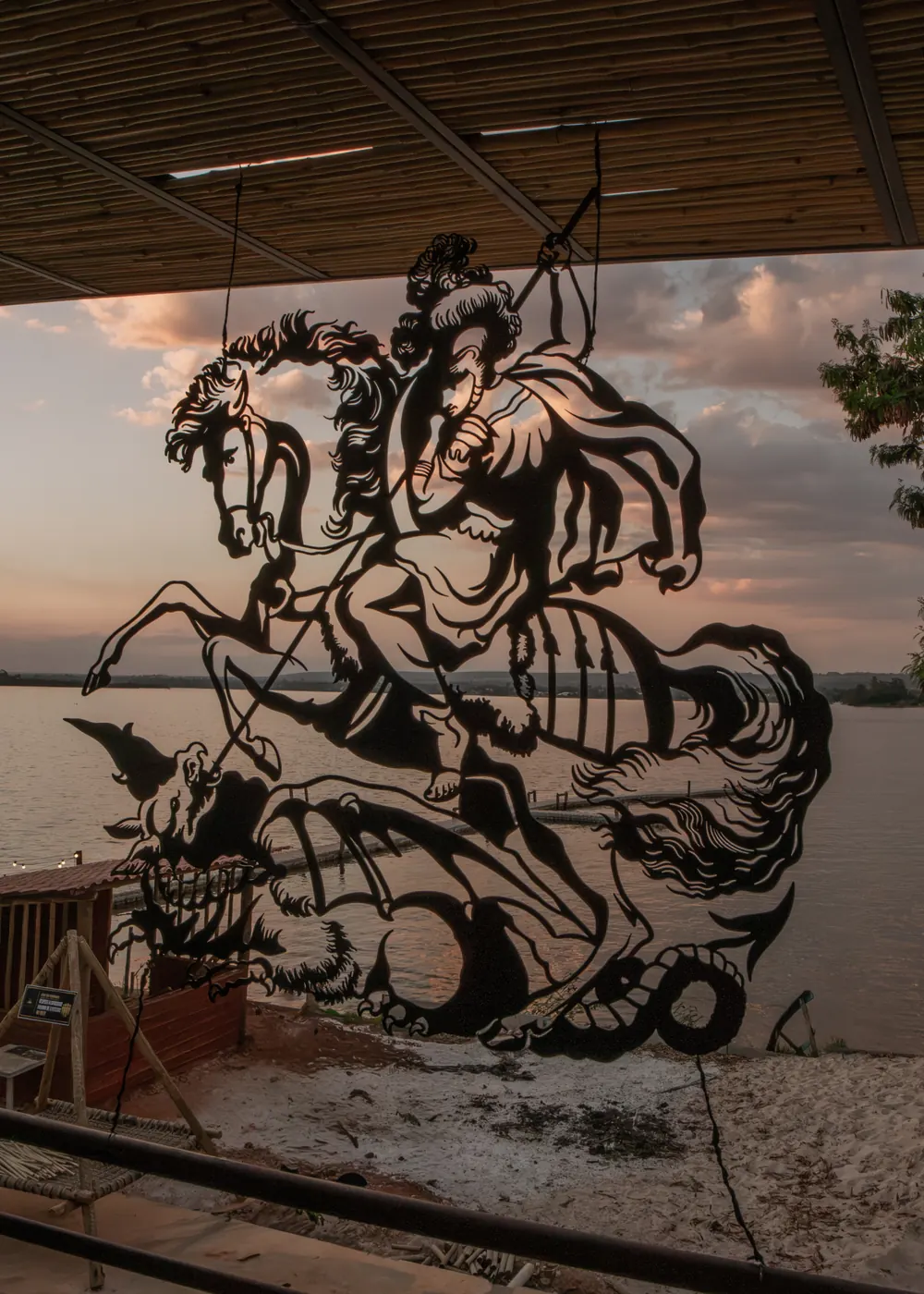Escultura vazada de São Jorge no Cápsula Bar, com o Lago Paranoá ao entardecer ao fundo