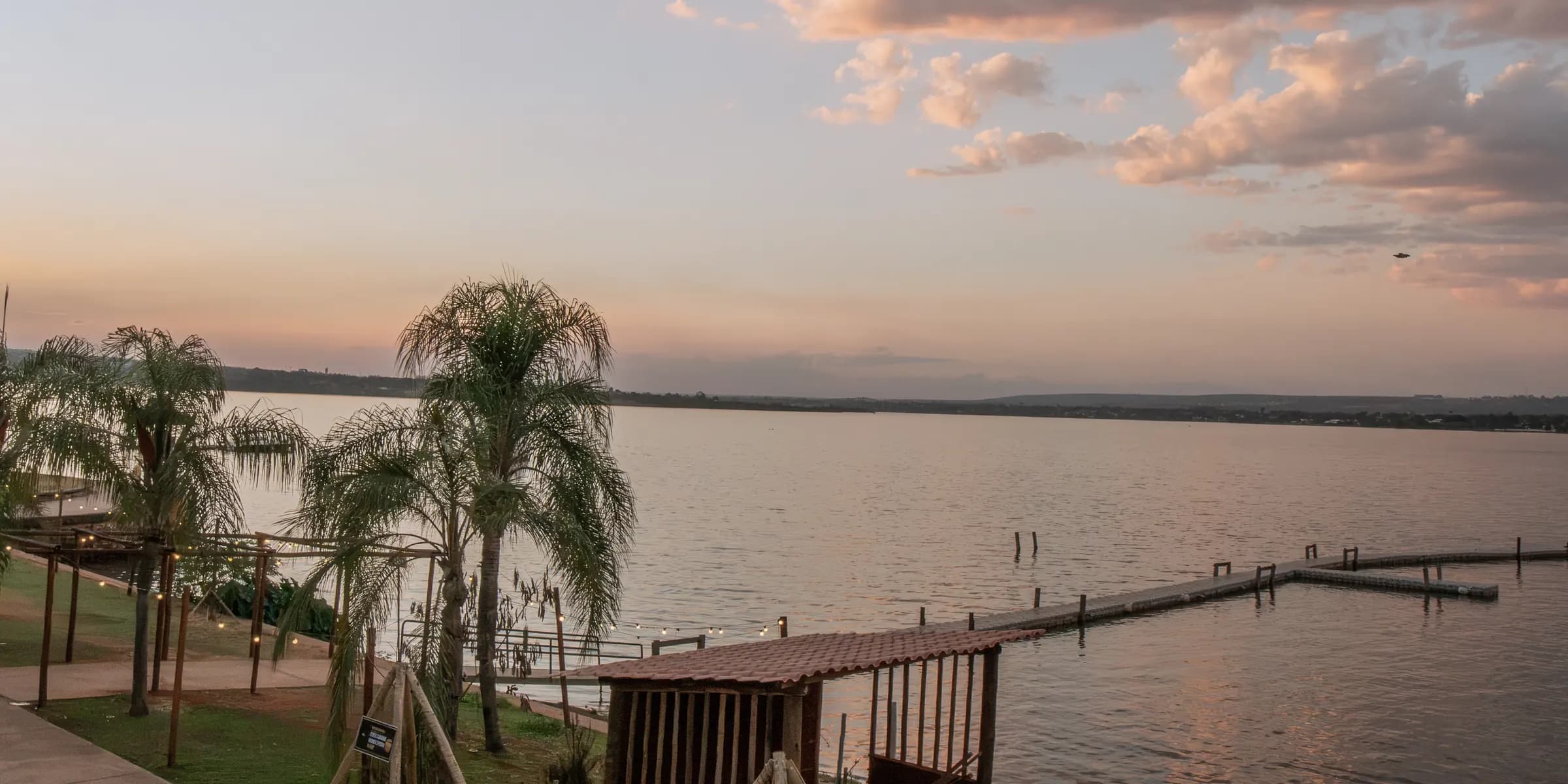 Cápsula Bar às margens do Lago Paranoá ao pôr do sol, com palmeiras e o píer sobre a água
