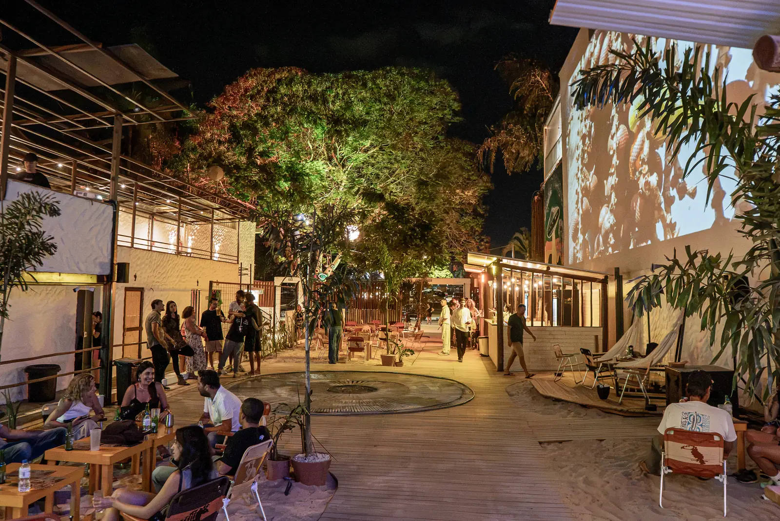 Pessoas reunidas no pátio de areia do Cápsula Bar à noite, entre árvores iluminadas