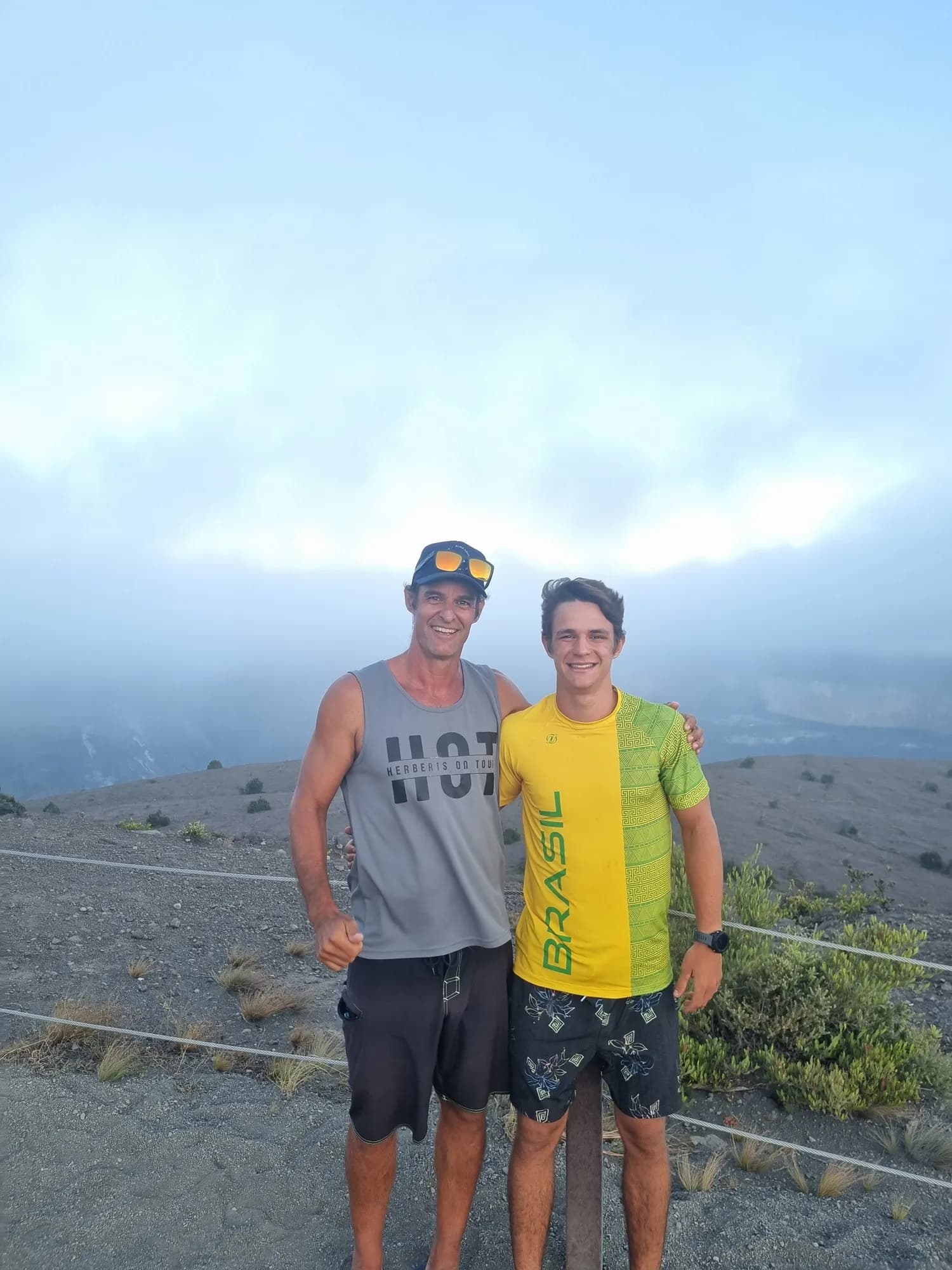 Marcelo e Rudah Bosi, pai e filho, abraçados na borda de uma cratera vulcânica coberta de névoa