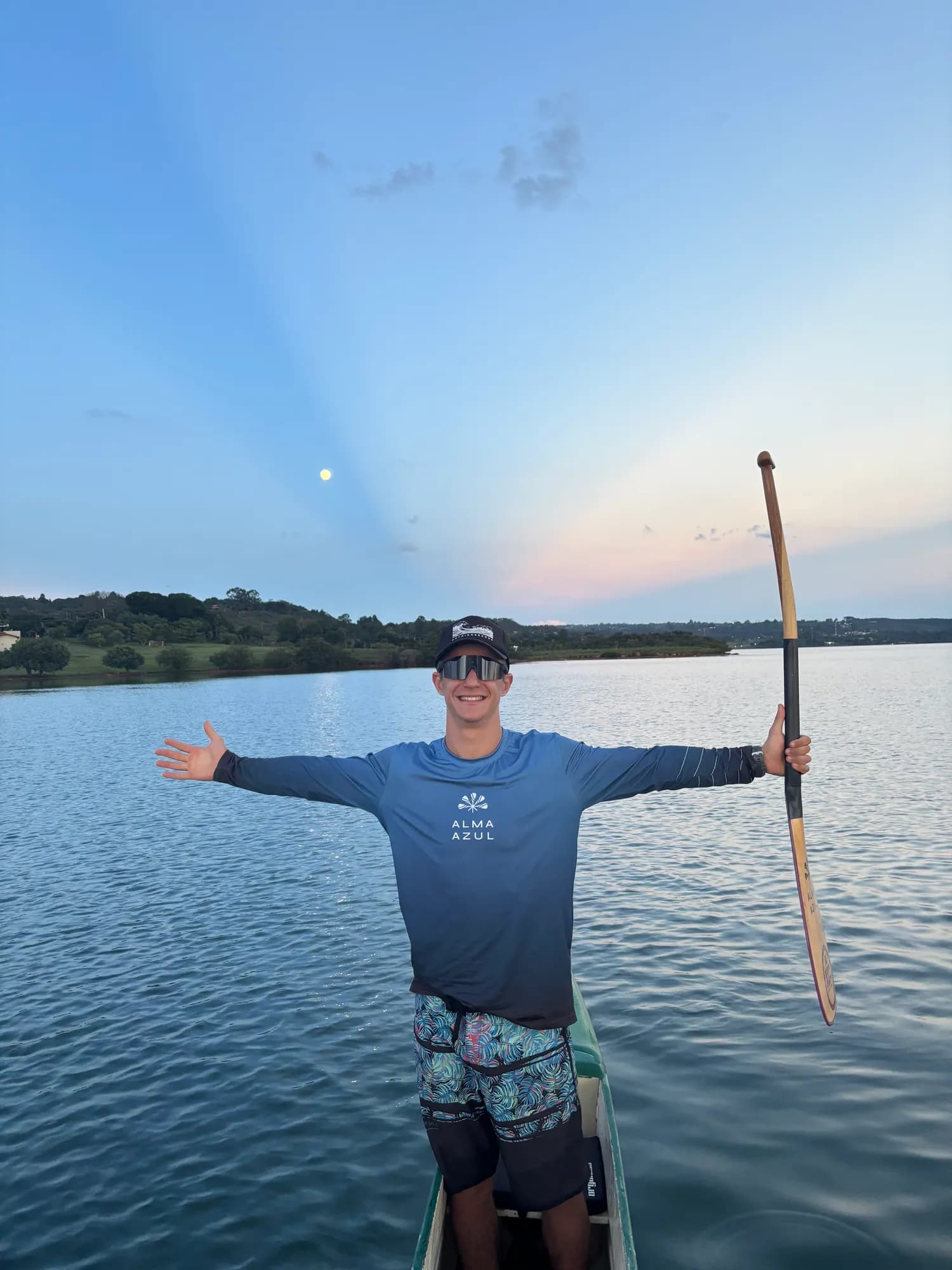 Rudah Bosi de braços abertos em uma canoa no Lago Paranoá ao entardecer, segurando um remo